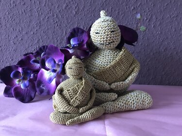 Zwei gehäkelte beige Buddha-Figuren, eine größer und eine kleiner, vor violetten Orchideen auf rosa Stoff