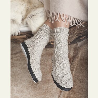 Person trägt cremefarbene, gestrickte Haussocken mit Zopfmuster und dunkler rutschfester Sohle.