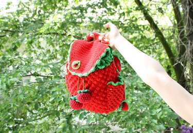 Red crocheted backpack with green ruffled trim and two berry pom-pom drawstrings held by a hand against green leaves