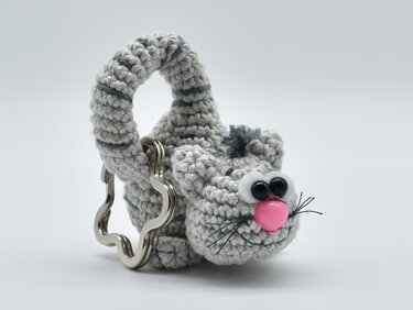 Gehäkelter grauer Katzen-Schlüsselanhänger mit rosa Nase und schwarzen Knopfaugen an einem wellenförmigen Metallring.