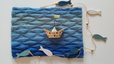 Gestrickter blauer Loop mit wellenförmigem Muster, Papierboot und Holzfischen