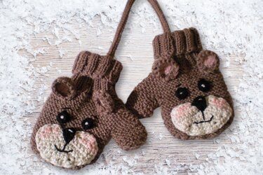 Braune gestrickte Baby-Fäustlinge mit Bärenmotiv und Verbindungsband auf schneebedecktem Holz