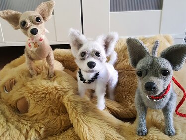 Drei gehäkelte Amigurumi-Hunde in Beige, Weiß und Grau mit Halsbändern auf flauschiger Decke