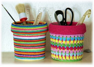 Zwei kompakte gehäkelte Utensilos mit umgeschlagenem Rand, eines gestreift, eines mit rosa Rand, gefüllt mit Schere, Lineal und Pinseln