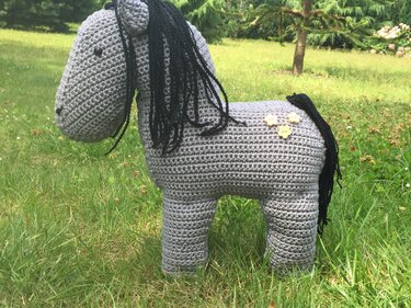 Gehäkeltes graues Pferd mit schwarzer Mähne und drei Blumenknöpfen auf Gras.
