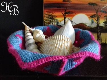 Gehäkeltes rundes Körbchen in Blau und Pink, gefüllt mit verschiedenen Muscheln, auf dunklem Untergrund vor einem Landschaftsgemälde