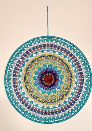 Rundes gehäkeltes Mandala in Blau-, Türkis-, Grün-, Gelb- und Rottönen an heller Wand.