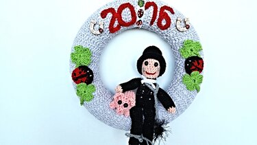 Runder, gehäkelter grauer Kranz geschmückt mit roten Zahlen '20:26', grünen Kleeblättern, roten Marienkäfern, Hufeisen und einem Schornsteinfeger, der ein rosa Schweinchen hält.