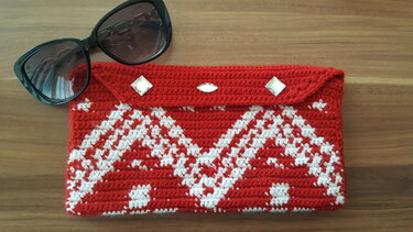 Rot-weiße, handgehäkelte Clutch mit Rautenmuster und drei funkelnden Strasssteinen auf dem Überschlag, liegend auf einer Holzoberfläche neben einer schwarzen Sonnenbrille.