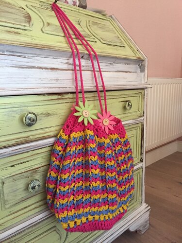 Gehäkelte Umhängetasche in Pink, Gelb und Blau mit Blumenknöpfen, hängt an einer grünen Kommode.