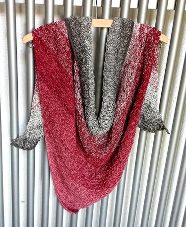 Gestricktes Dreieckstuch in Rot-, Rosa- und Grautönen auf einem Holzkleiderbügel vor Metallrohren hängend.