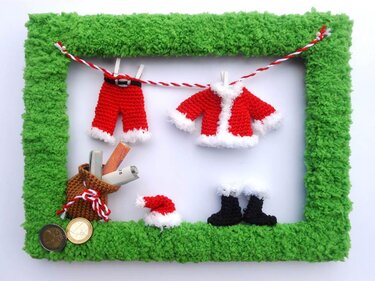 Gehäkelte Mini-Nikolausjacke, Hose, Mütze und Stiefel an Schnur in grünem Rahmen, daneben kleiner Sack mit Münzen