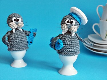 Zwei gehäkelte Walross-Eierwärmer auf Eierbechern, einer mit Seemannsmütze, beide mit kleinem blauen Fisch, vor blauem Hintergrund.
