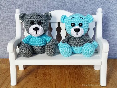 Zwei gehäkelte Amigurumi-Teddybären (grau und türkis) auf einer weißen Miniaturbank vor neutralem Hintergrund.