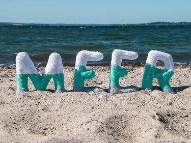 Gehäkelte Buchstaben 'MEER' in Weiß, Türkis und Grau auf Sandstrand vor dem Meer