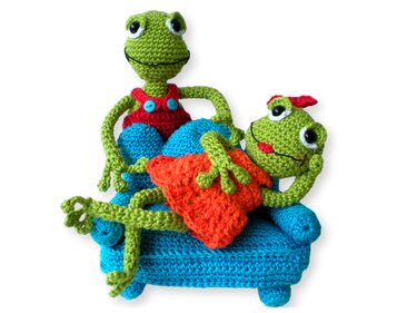 Zwei gehäkelte grüne Frösche auf einem kleinen blauen Sofa, einer mit roter Latzhose, der andere mit orangefarbenem Kissen.