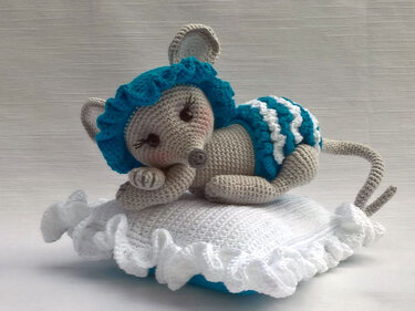 Beige crocheted amigurumi mouse with teal ruffled bonnet and diaper lying on a white ruffled pillow