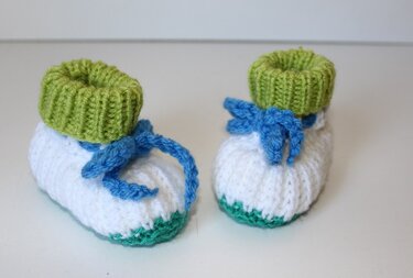 Paar gestrickter Babyschuhe weiß mit grünem Umschlag und blauer Kordel