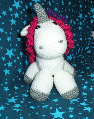 Weißes gehäkeltes Einhorn-Amigurumi mit pink-lila Lockenmähne, grauen Hufen und grauem Horn auf blauem Sternenstoff