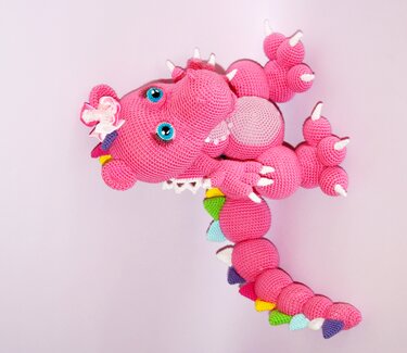 Pink gehäkelter Amigurumi-Drache mit blauen Augen und bunten Rückenzacken