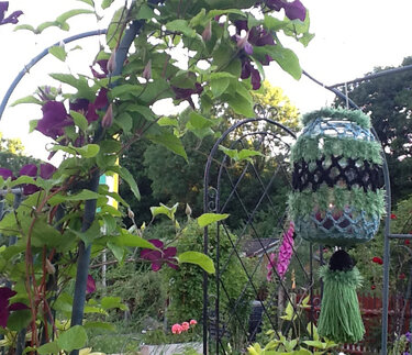 Gehäkelte Laternenhülle mit Quaste hängt am Metallbogen neben Kletterpflanze mit violetten Blüten