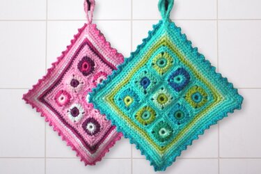 Zwei gehäkelte Granny-Square-Topflappen in Pink und Türkis hängen an einer gefliesten Wand.
