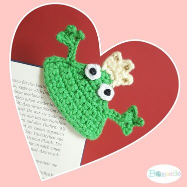 Gehäkeltes grünes Frosch-Lesezeichen mit cremefarbener Krone auf einer Buchseite