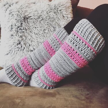 Nahaufnahme von Beinen mit grau-pink gestreiften gehäkelten Beinstulpen auf einem braunen Sofa mit flauschigem Kissen im Hintergrund.