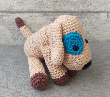 Beiger gehäkelter Amigurumi-Hund mit blauem Augenring, braunen Pfoten und schwarzer Nase