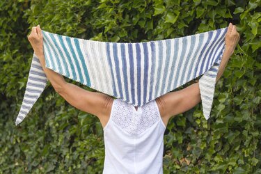 Person hält blau-weiß gestreiftes gestricktes Dreieckstuch vor grüner Hecke