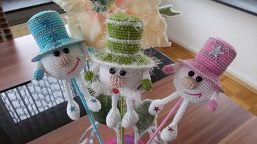 Drei gehäkelte, schneemannähnliche Figuren mit blauen, grünen und rosa Zylinderhüten auf Stäben auf einer Holzoberfläche.