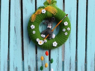 Gehäkelter grüner Osterkranz mit grauem Hasen, Pinsel, weißen Blüten und bunten gehäkelten Eiern vor hellblauer Holzwand.