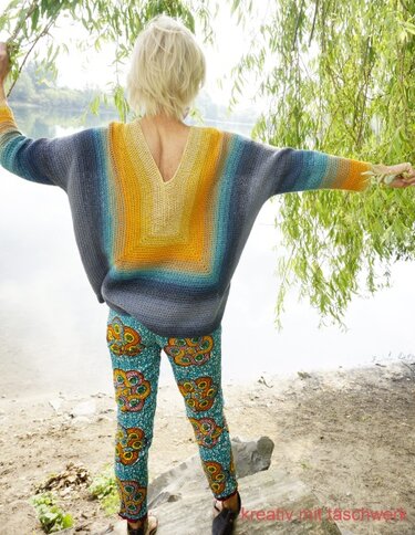 Rückansicht einer Frau in einem gehäkelten Oversize-Pullover mit Farbverlauf und V-Ausschnitt im Rücken, stehend am Ufer unter Weidenzweigen.