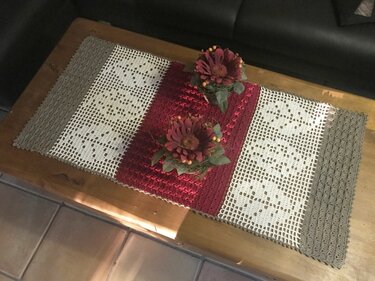 Gehäkelter Tischläufer in Beige, Weiß und Bordeaux mit Filet-Herz- und Blumenmotiven auf einem Holztisch, dekoriert mit zwei künstlichen Blumenarrangements