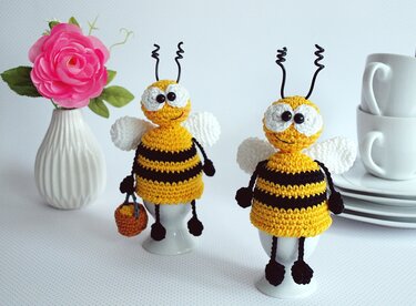 Zwei gehäkelte Bienen-Eierwärmer in Gelb und Schwarz mit weißen Flügeln und gewellten Fühlern auf Eierbechern, eine mit kleinem Korb.
