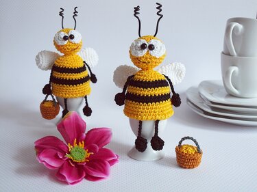 Zwei gehäkelte Bienen-Eierwärmer in Gelb und Schwarz auf Eierbechern, davor eine pinke Blüte und ein kleines gehäkeltes Körbchen, im Hintergrund gestapelte Tassen und Teller