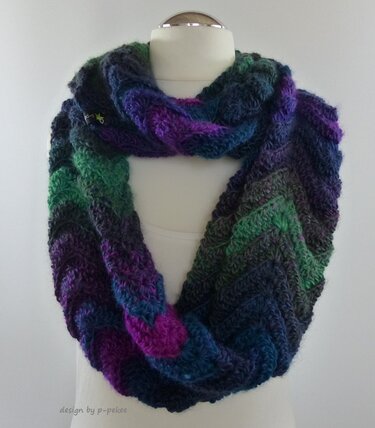 Gehäkelter Loop-Schal mit Chevron-Muster in Blau-, Violett- und Grünverläufen an einer weißen Schaufensterpuppe.