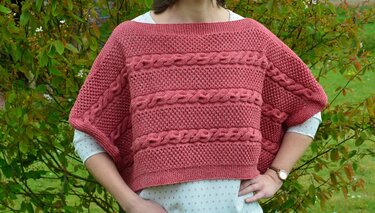 Eine Person trägt einen korallfarbenen Strickponcho mit Zopf- und Lochmustern vor grünem Hintergrund.