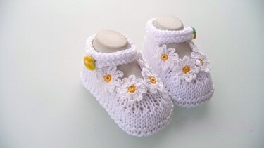Zwei weiße, gestrickte Babyschuhe mit Margeriten-Applikationen und gelben Knöpfen auf hellem Hintergrund.