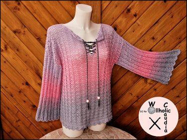 Gehäkelter Damenpullover in Rosa‑Lila‑Grau mit Schnür‑V‑Ausschnitt und Muschelkante, präsentiert auf Schaufensterpuppe vor Holzpaneelen.