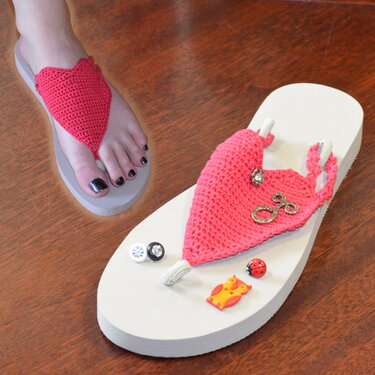 Rote Flip-Flops mit gehäkeltem Herzriemen; ein Schuh wird getragen, der andere mit Knöpfen und Charms auf Holz.