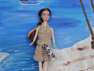Puppe in beigem gehäkeltem Neckholder-Top und Shorts mit gemusterter Schärpe vor einer gemalten Strand- und Meer-Kulisse