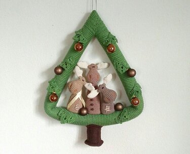 Gehäkelter grüner Weihnachtsbaum an weißer Wand, mit drei beigebraunen gehäkelten Hasenfiguren, kupferfarbenen Kugeln und braunem Stamm.