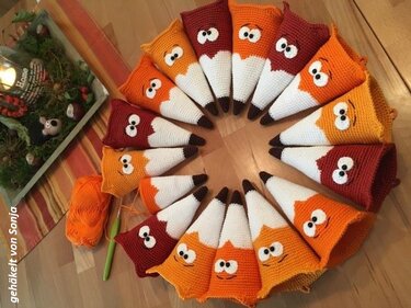 Kreis aus gehäkelten, kegelförmigen Wichteln in Rot und Orange mit weißen Spitzen; Garn und Häkelnadel auf Holzuntergrund.