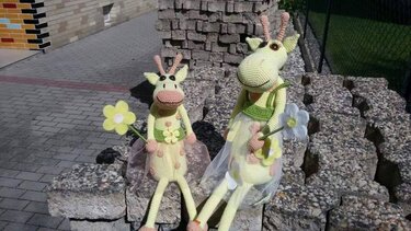 Zwei handgehäkelte Amigurumi‑Giraffen in Pastellgelb mit rosa Akzenten sitzen auf einer Steinmauer und halten gehäkelte Blumen.