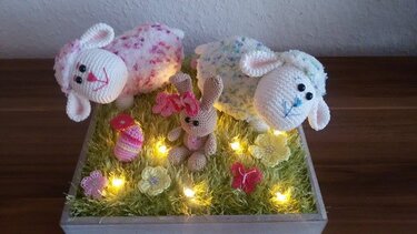 Zwei gehäkelte Lämmer und ein Häschen in einer Dekobox mit Gras, Blumen und Lichterkette