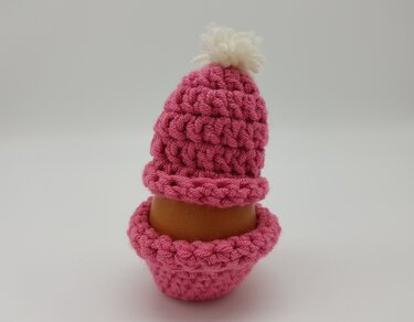 Braunes Ei im pinken gehäkelten Eierwärmer mit weißem Bommel vor hellem Hintergrund.