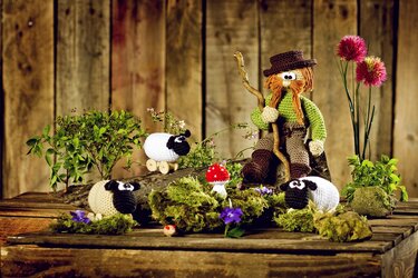 Gehäkelte Bauernfigur mit Stock und drei kleinen gehäkelten Schafen auf Holzuntergrund mit Moos und Blumen