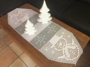 Weißer gehäkelter Tischläufer mit Rentiermotiv und zwei weißen Tannenbaumfiguren auf Holztisch
