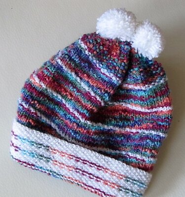 Gestrickte Babymütze mit bunten Streifen, weißem Umschlag und zwei weißen Bommeln.